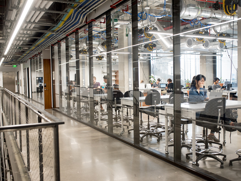 Close up of concrete hallway with glass windows with students working at worktables